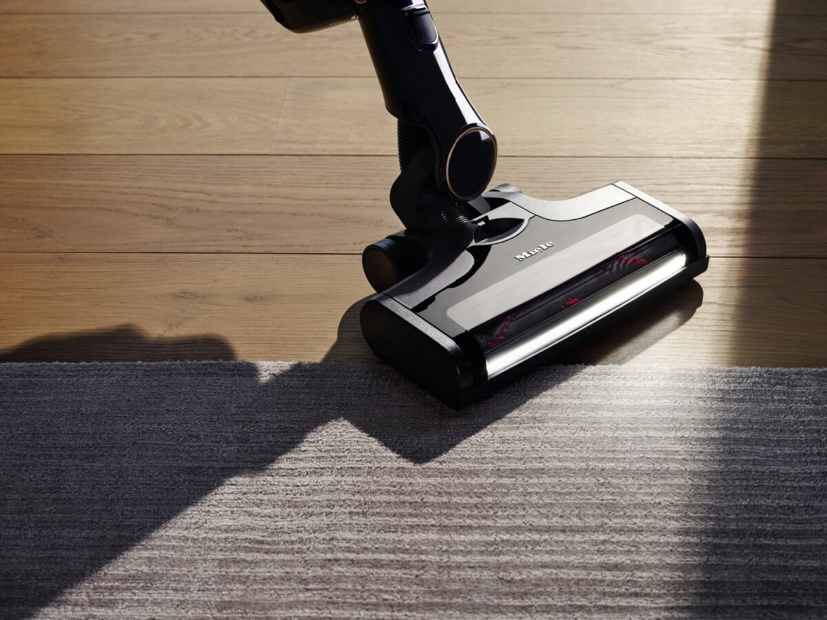 Zbliżenie czarnej głowicy odkurzacza na dywanie. Podłoga jest drewniana. Cień na dywanie.