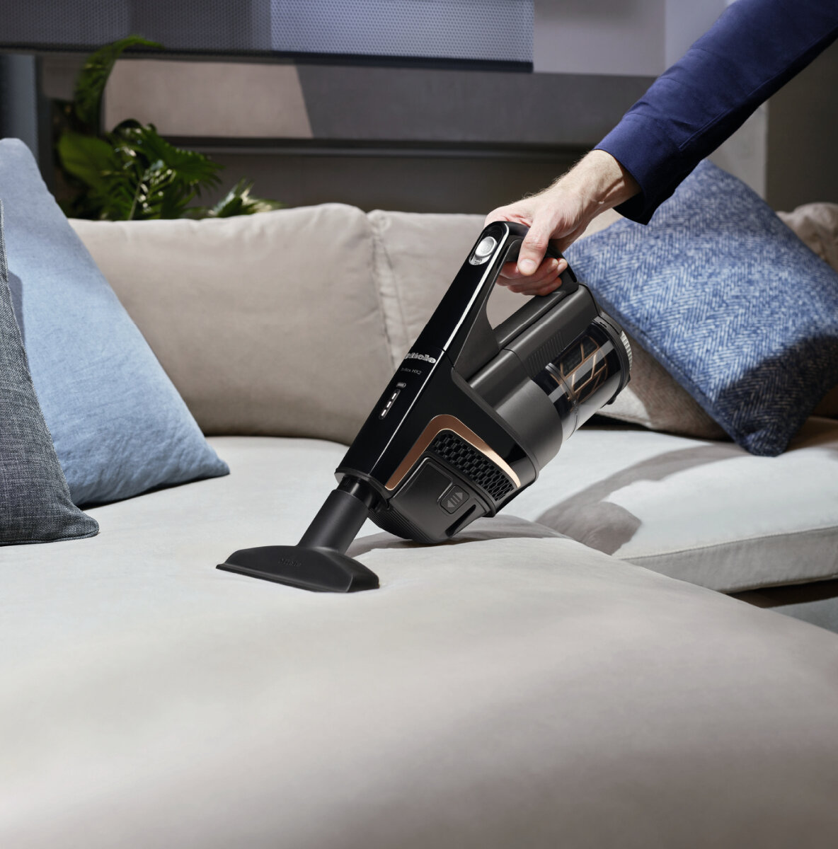 Ręka odkurza sofę. Odkurzacz jest czarny i złoty, na jasnoszarej sofie. Niebieskie poduszki w tle.