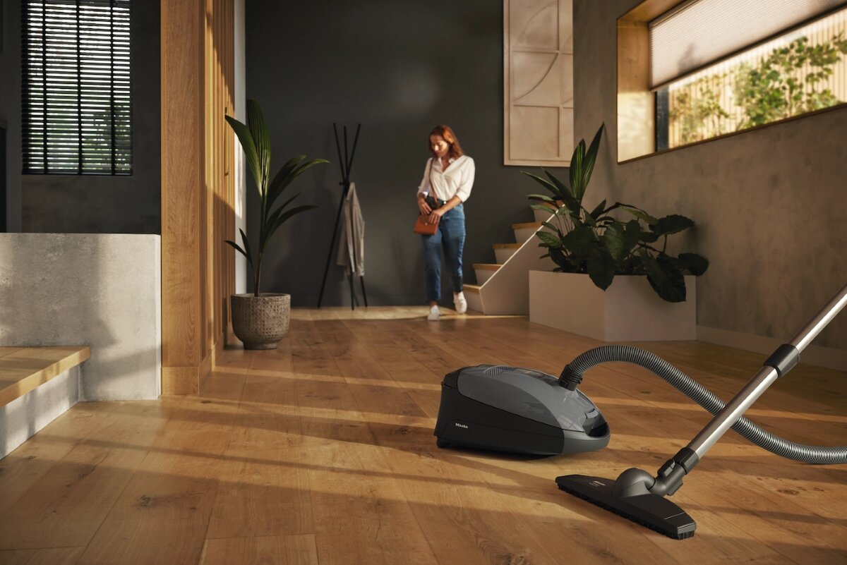 Odkurzacz stoi na drewnianej podłodze. Kobieta idzie w tle korytarza.