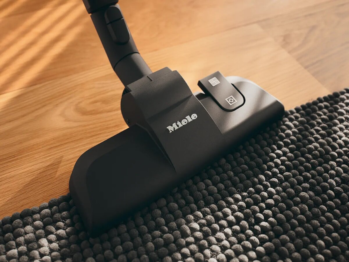 Głowica odkurzacza znajduje się na szarym, teksturowanym dywanie. Słowo 'Miele' znajduje się na głowicy odkurzacza.