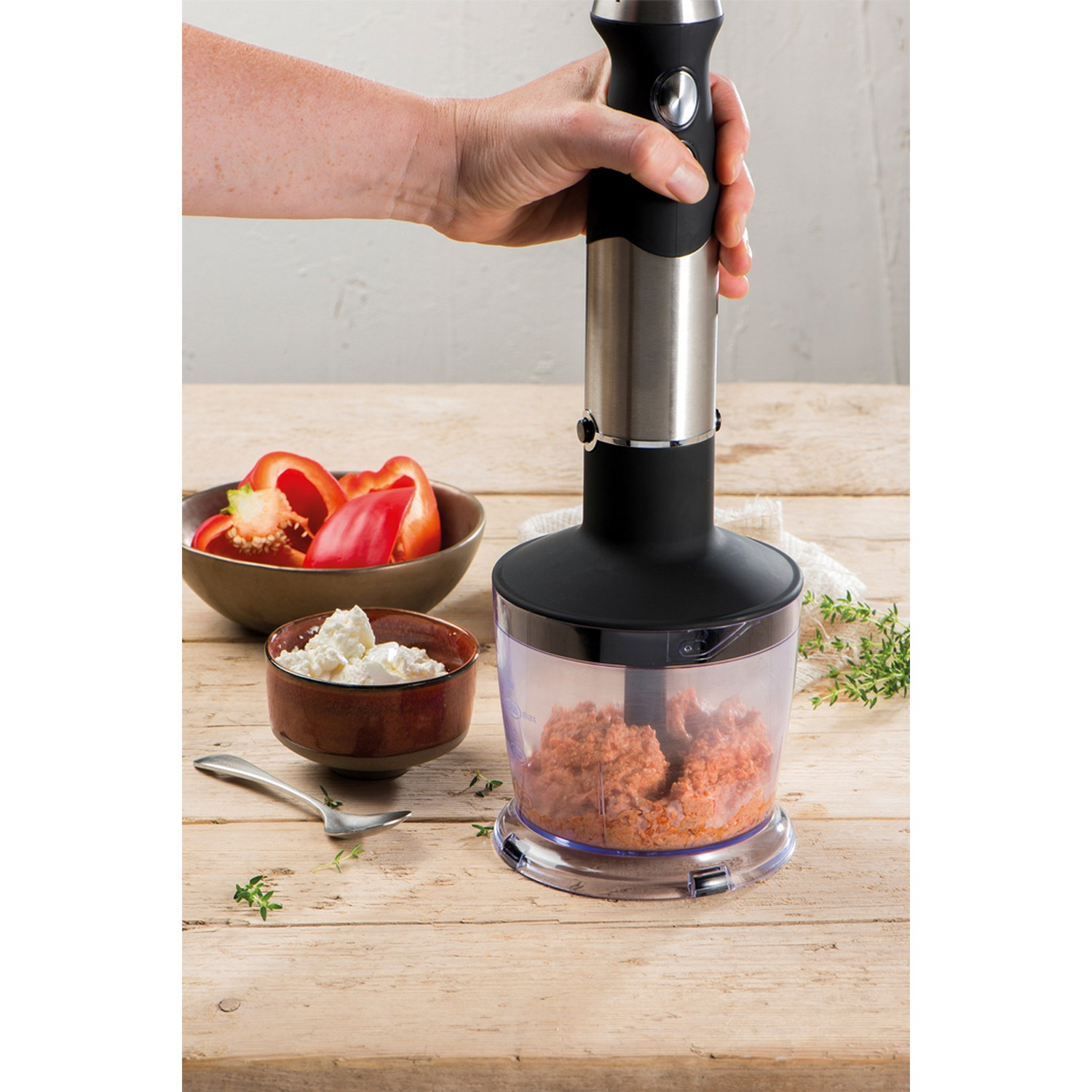 Hand die een blender vasthoudt met voedsel in de container, met ingrediënten op tafel.