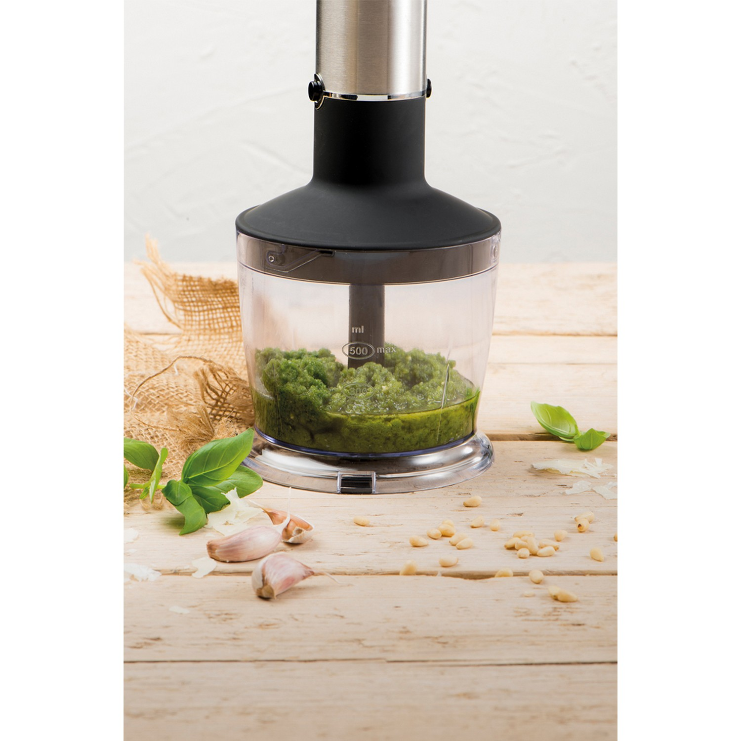 Blender met pesto erin, omringd door ingrediënten zoals basilicum, pijnboompitten en knoflook.