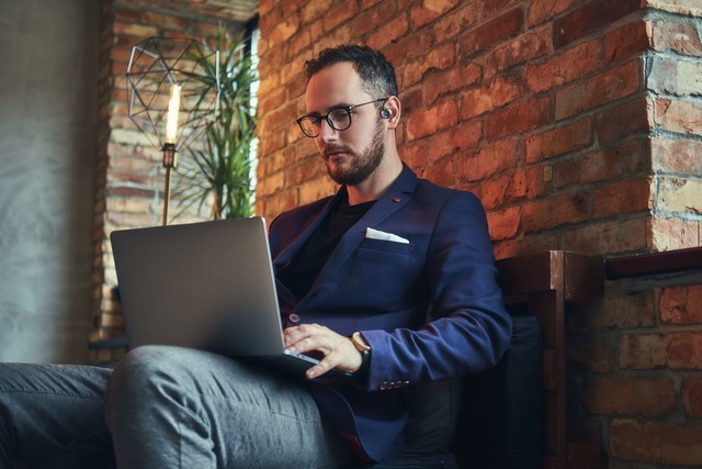 Hombre con gafas y blazer, trabajando en portátil, sentado junto a pared de ladrillo y lámpara.