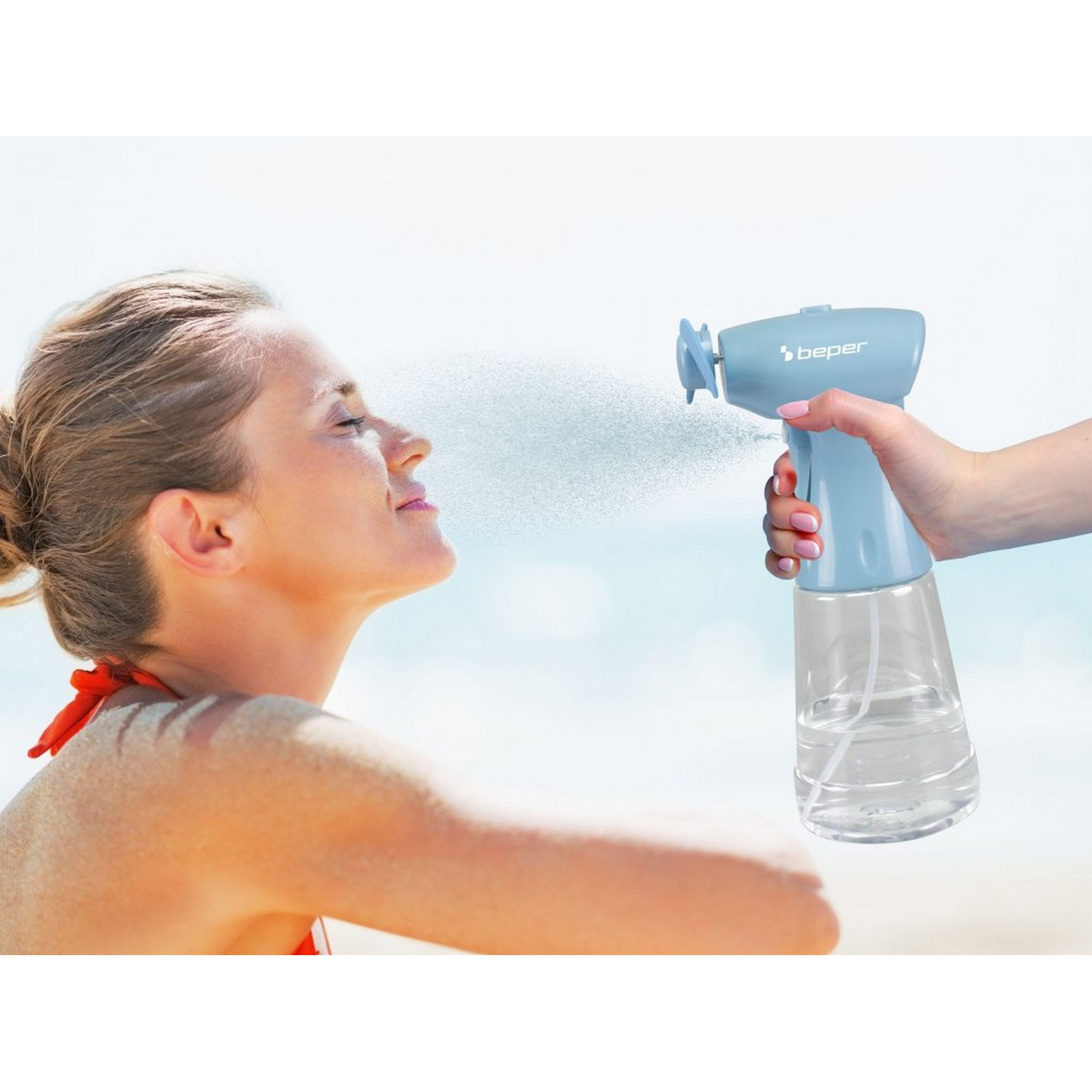 Visage d'une femme aspergé d'eau par un appareil bleu. Arrière-plan : plage et ciel bleu.