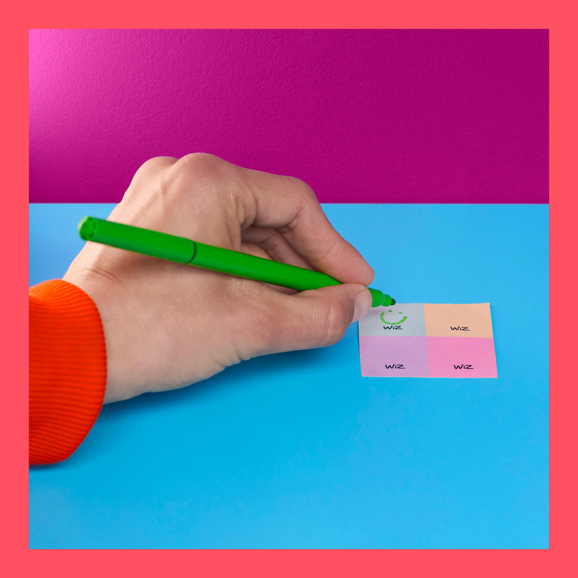 Hand zeichnet Smiley auf Haftzettel, vor blau-rosa Hintergrund.