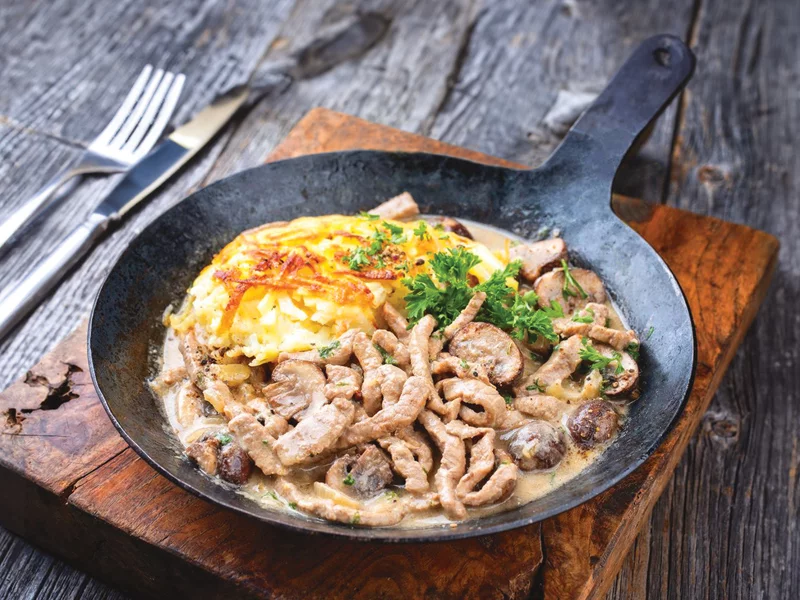Piatto con cibo. Contiene carne, patate e funghi. Posizionato su una superficie di legno.