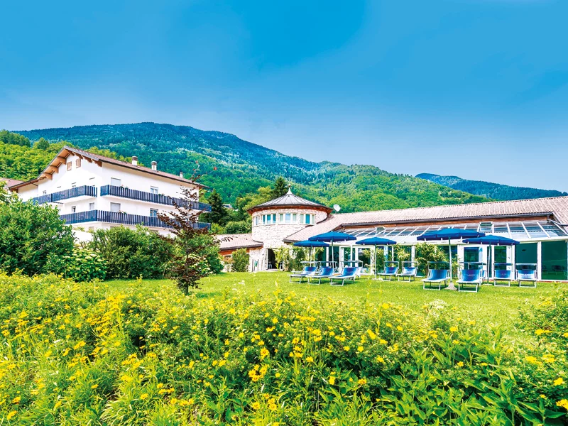 Vista panoramica di un hotel con prato verde, ombrelloni blu e montagne.