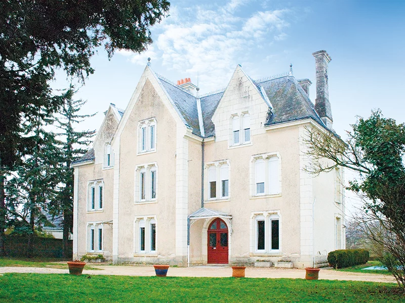 Bâtiment crème avec fenêtres, porte rouge et arbres en arrière-plan.