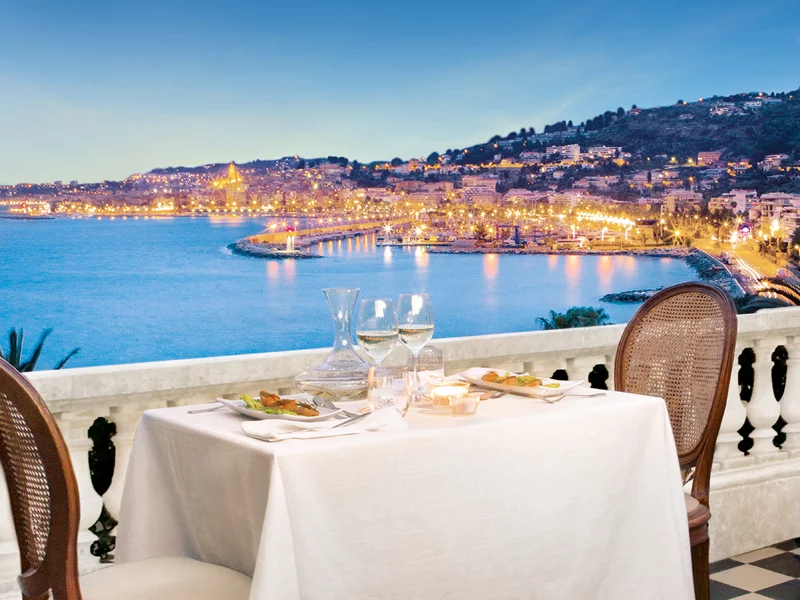 Table dressée pour dîner sur un balcon surplombant une ville au crépuscule, vue sur l'océan.