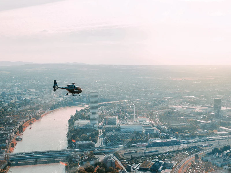 Elicottero in volo su città con fiume, edifici e cielo soleggiato.