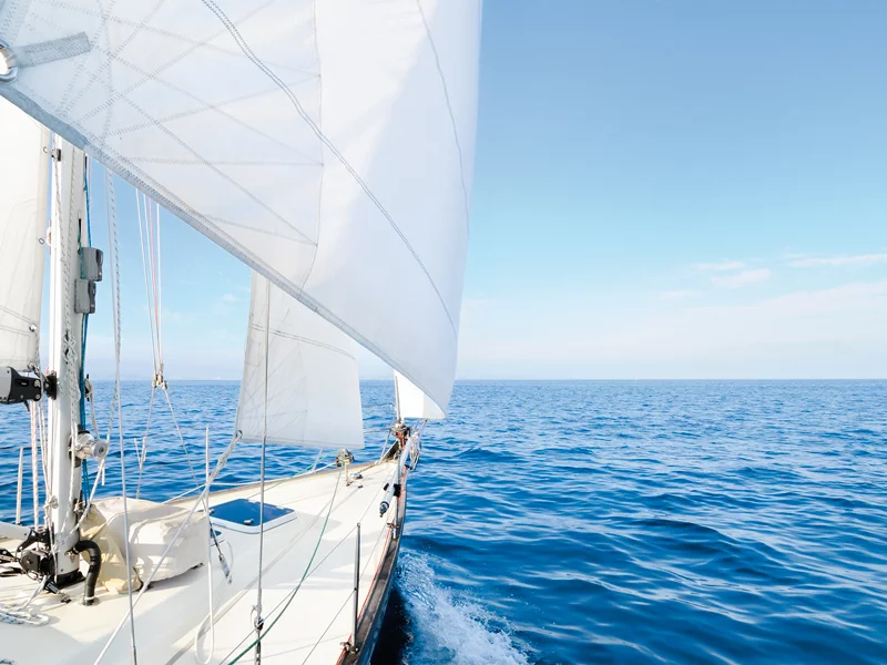Una barca a vela in mare, vele spiegate, cielo blu sopra. La barca si muove in avanti, creando acqua bianca.
