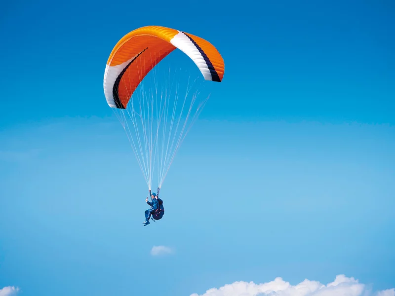 Una persona fa parapendio. Un paracadute arancione e bianco è sopra di loro. Cielo blu.