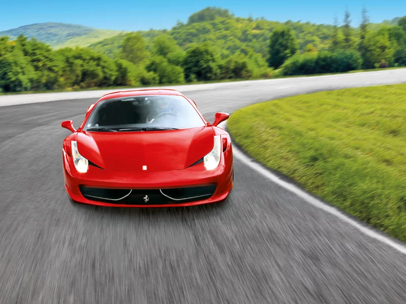 Un'auto sportiva Ferrari rossa sfreccia su una strada con colline verdi e cielo blu.