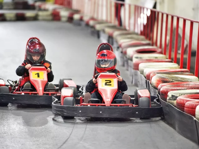 Zwei Kinder in roten Go-Karts auf einer Rennstrecke. Sie tragen Helme und schauen nach vorne. Rote und weiße Barrieren umgeben.