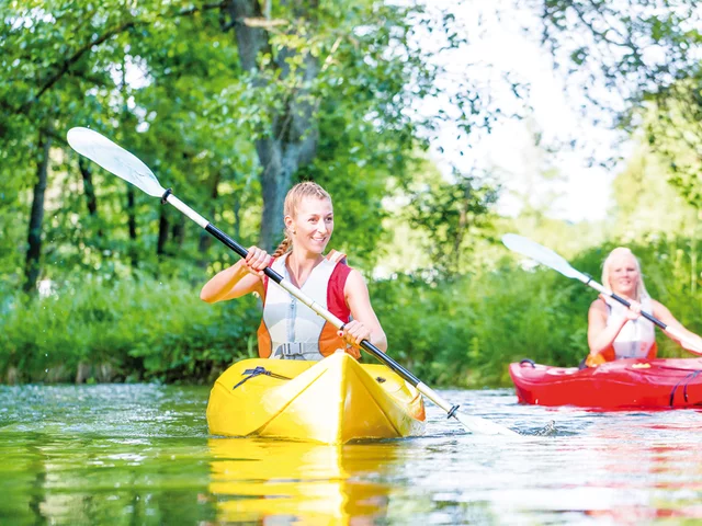 Zwei Frauen beim Kajakfahren auf einem Fluss mit grünen Bäumen im Hintergrund.