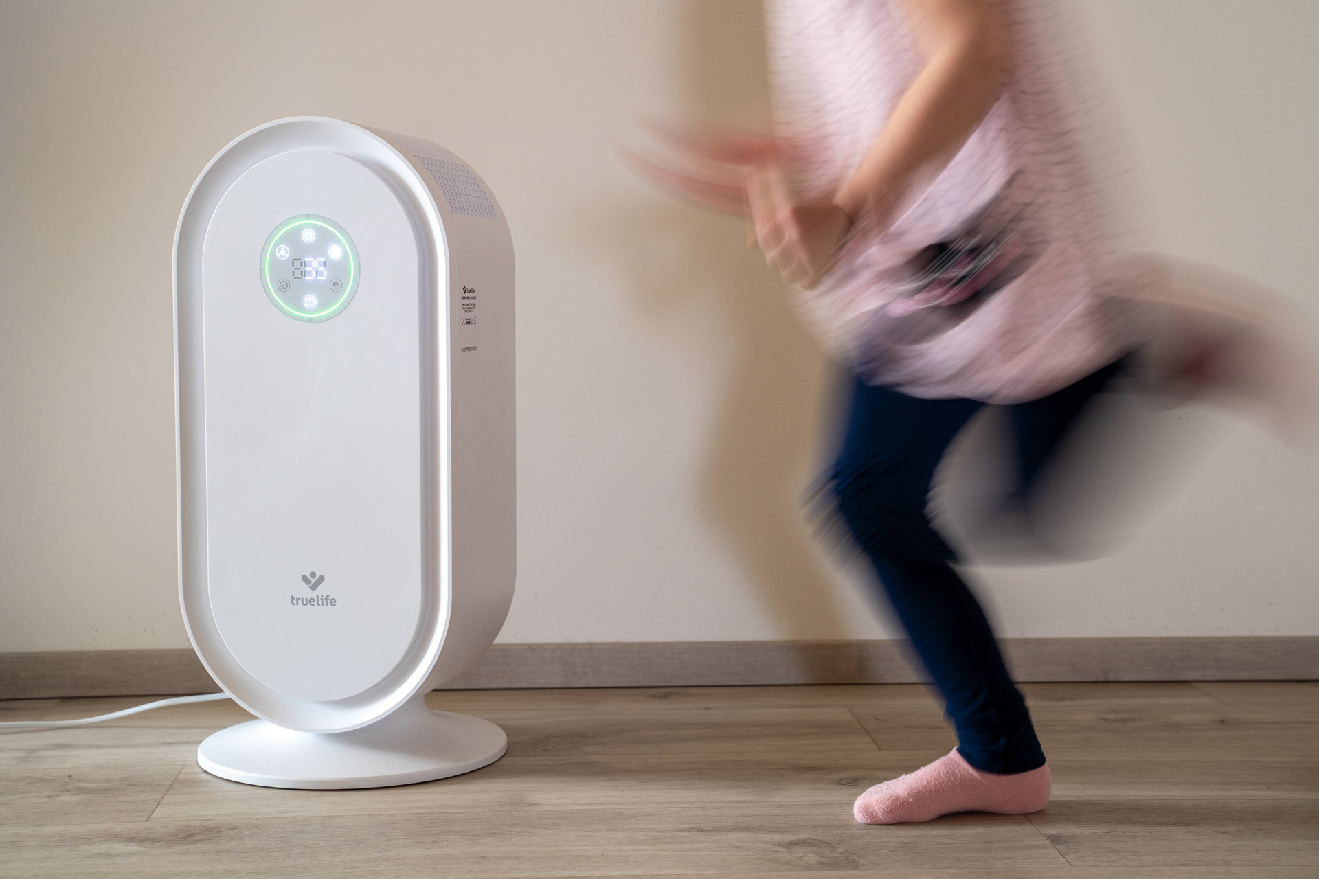 Egy fehér légtisztító digitális kijelzővel. Egy személy lábai elmosódottak, mellette futva.