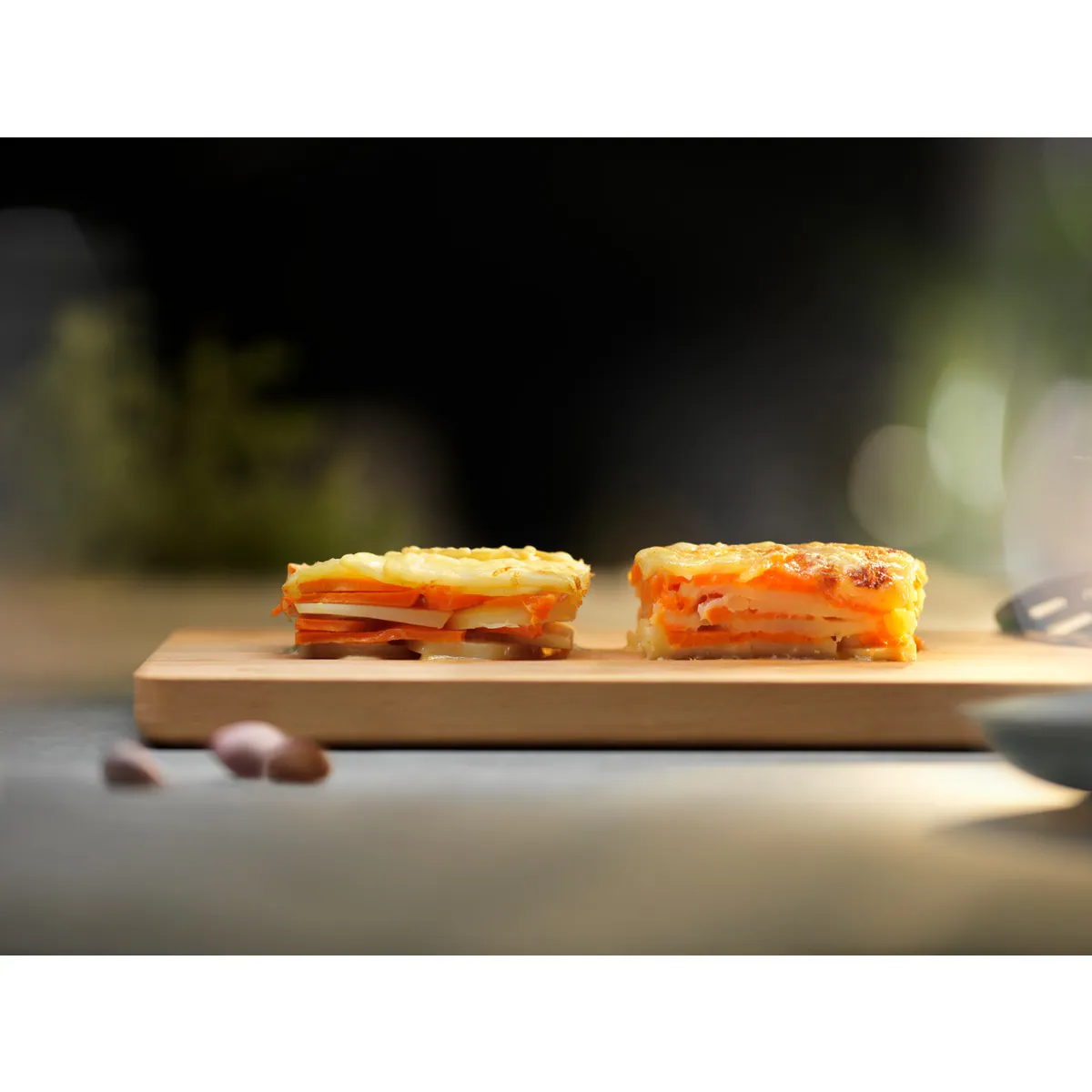 Dos porciones de un plato gratinado en una tabla de madera, fondo borroso con luces.