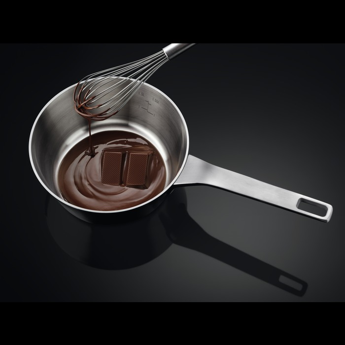 Een roestvrijstalen pan met gesmolten chocolade en een garde erin, op een zwart oppervlak.