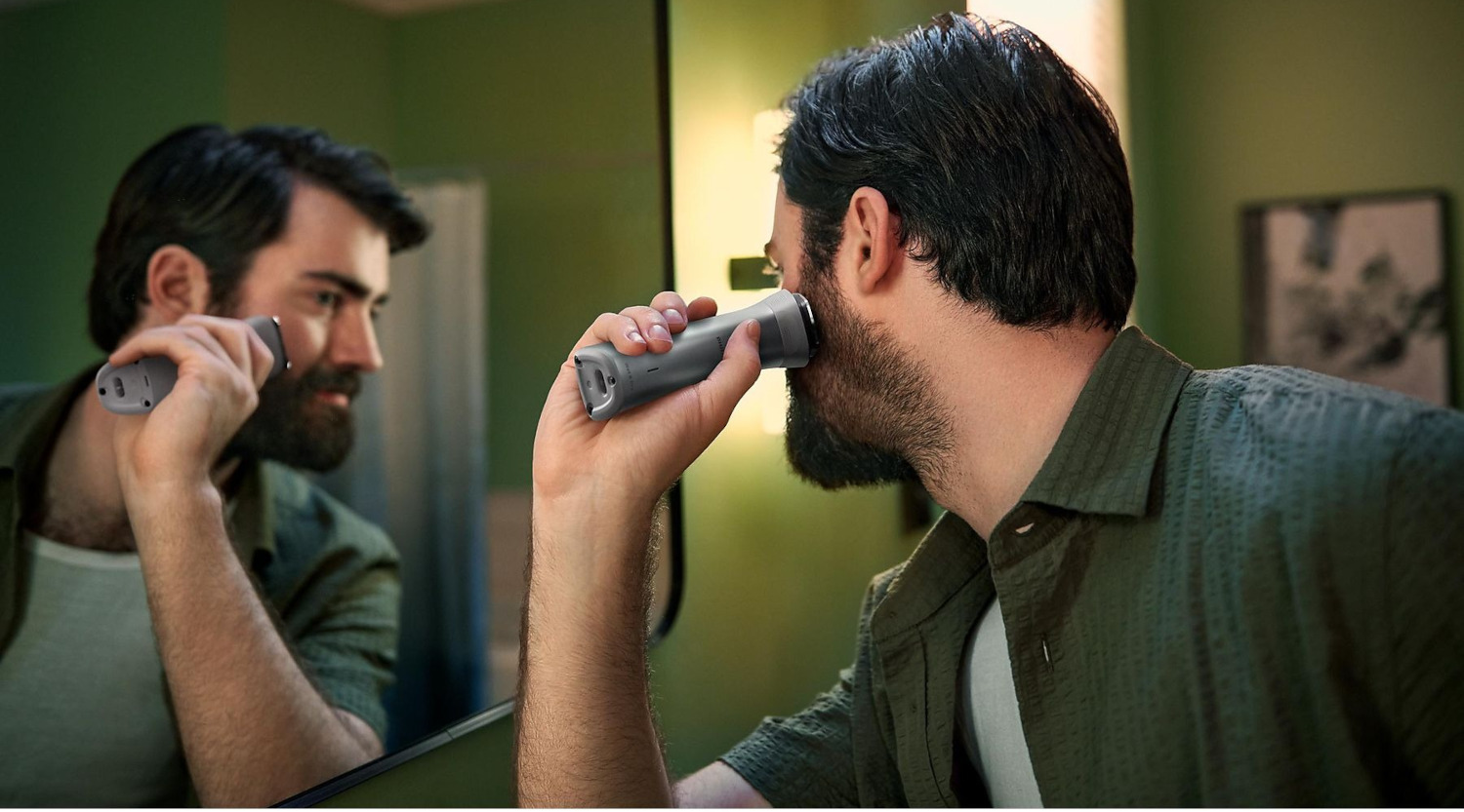Un homme se rase la barbe devant un miroir de salle de bain avec un rasoir électrique gris. Reflet visible.