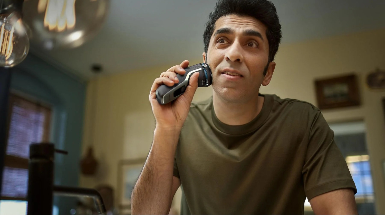 Man shaves his face with an electric shaver. He looks up with a surprised expression. Background is a mirror.