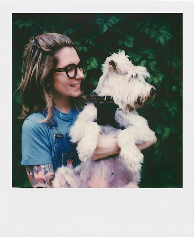 Mujer con gafas sosteniendo un perro blanco y esponjoso. Fondo de follaje verde.