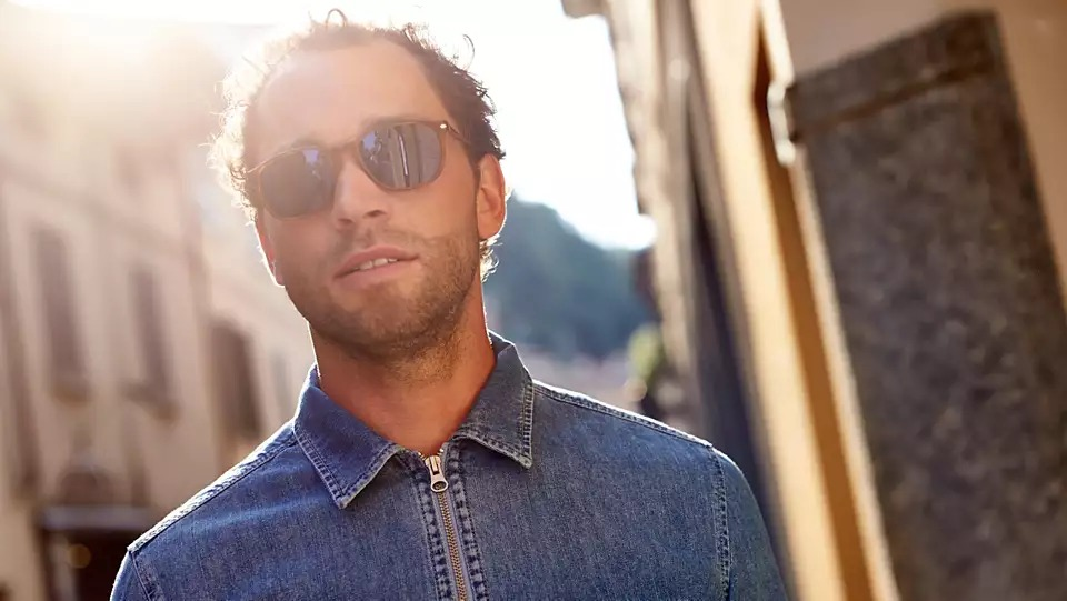 Man in sunglasses and denim jacket, posing outdoors in sunlight, with a blurred background.