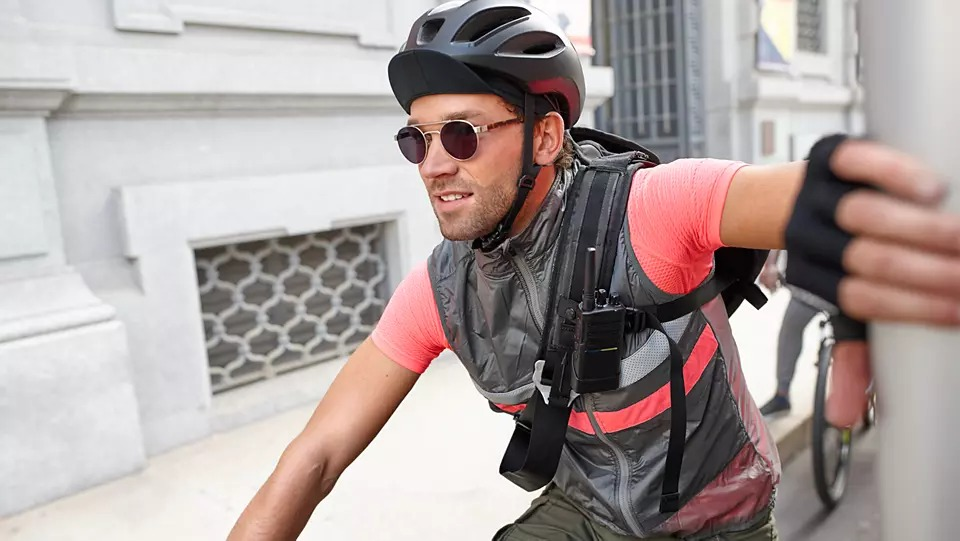 Cyclist riding on a city street. He is wearing sunglasses, a vest, and a helmet.