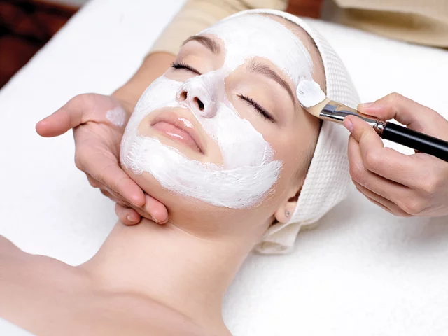 Il viso di una donna è coperto da una maschera bianca, con un pennello che la applica. È sdraiata.