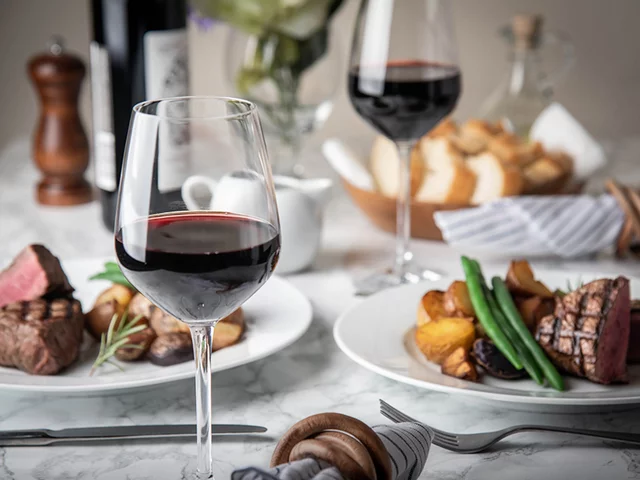 Due bicchieri di vino rosso con bistecca e verdure su un tavolo di marmo.