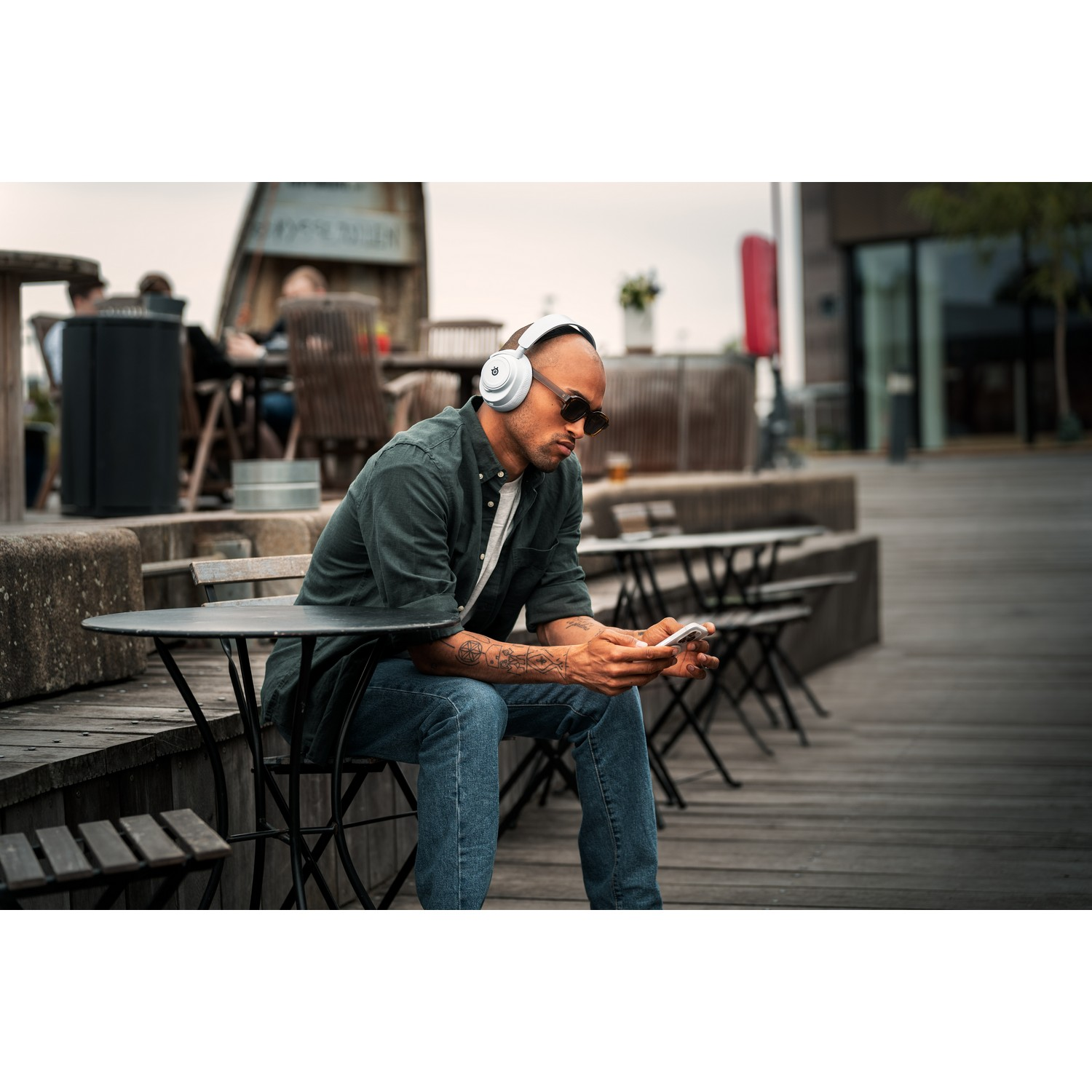 Man wearing sunglasses and headphones sits at a table, looking at his phone. Background shows other people.