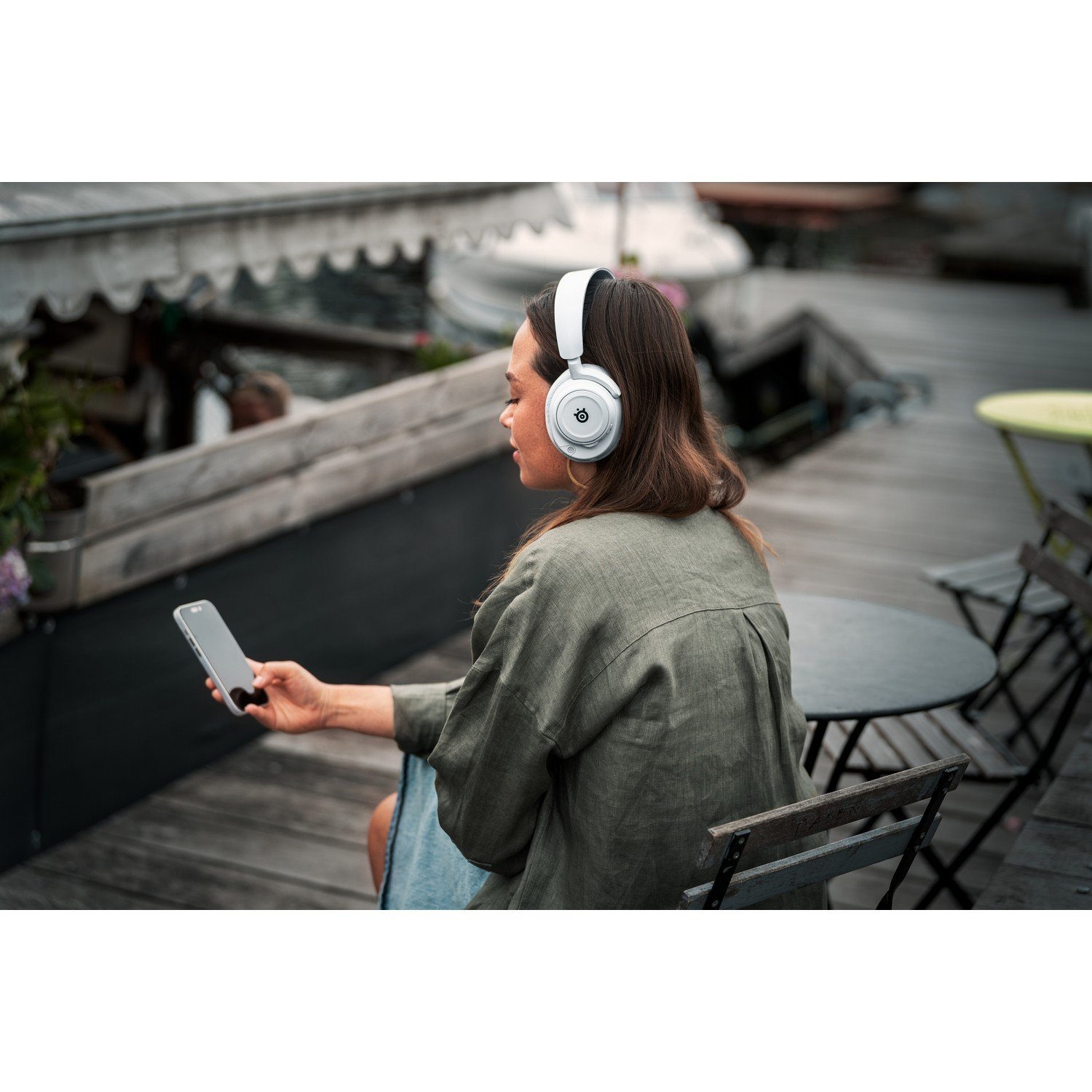 Woman with white headphones holds a phone, sitting outside. Background is a dock.