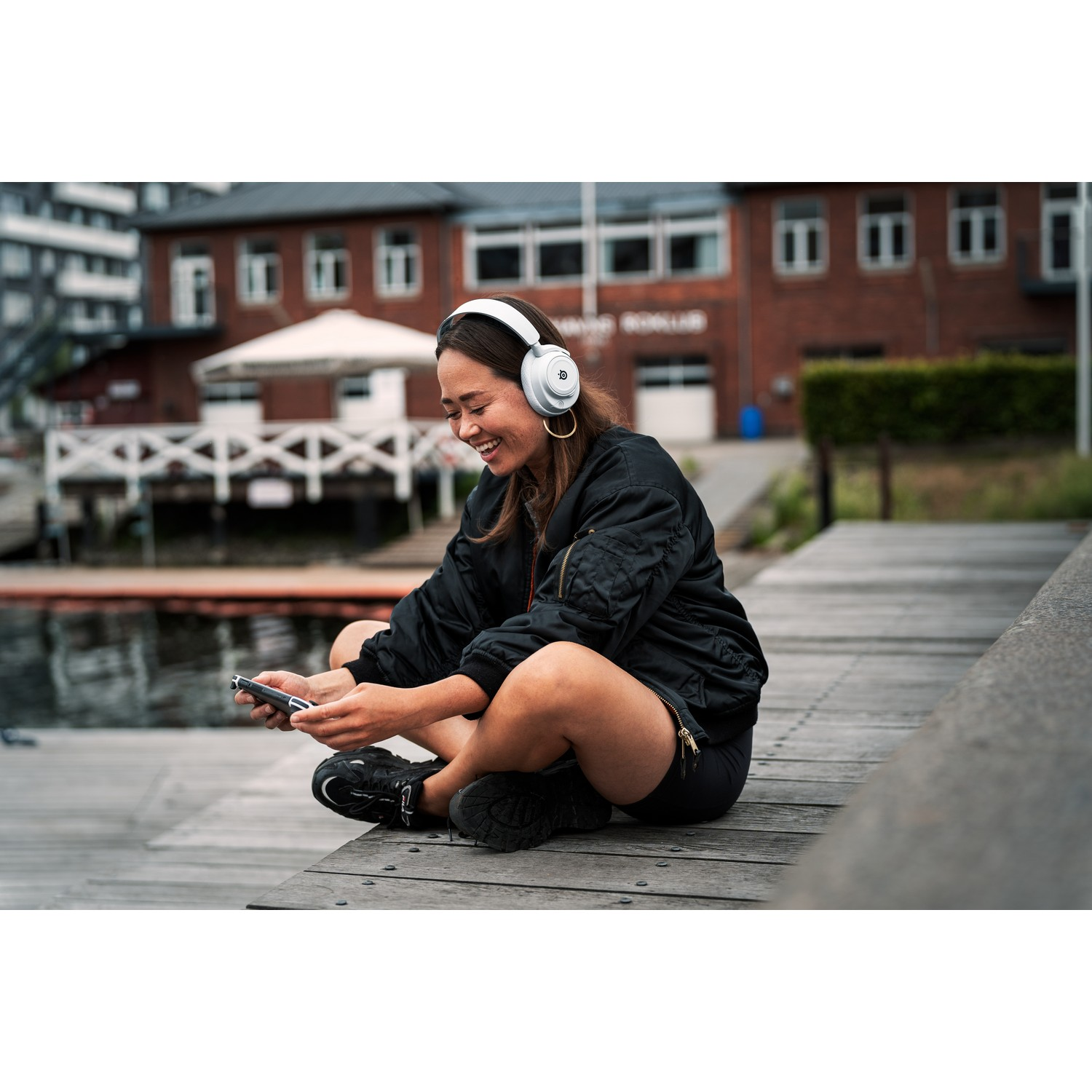 Woman sitting on a dock, listening to music with white headphones and smiling. Buildings in the background.