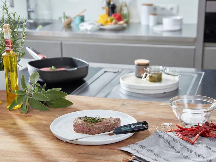 Steak auf einem Teller mit Thermometer, Kräutern, Öl, Salz und Bratpfanne im Hintergrund.