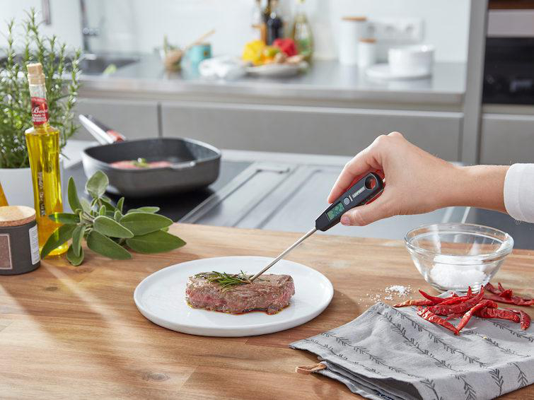Eine Hand verwendet ein Thermometer, um ein Steak auf einem weißen Teller zu prüfen, umgeben von Zutaten.