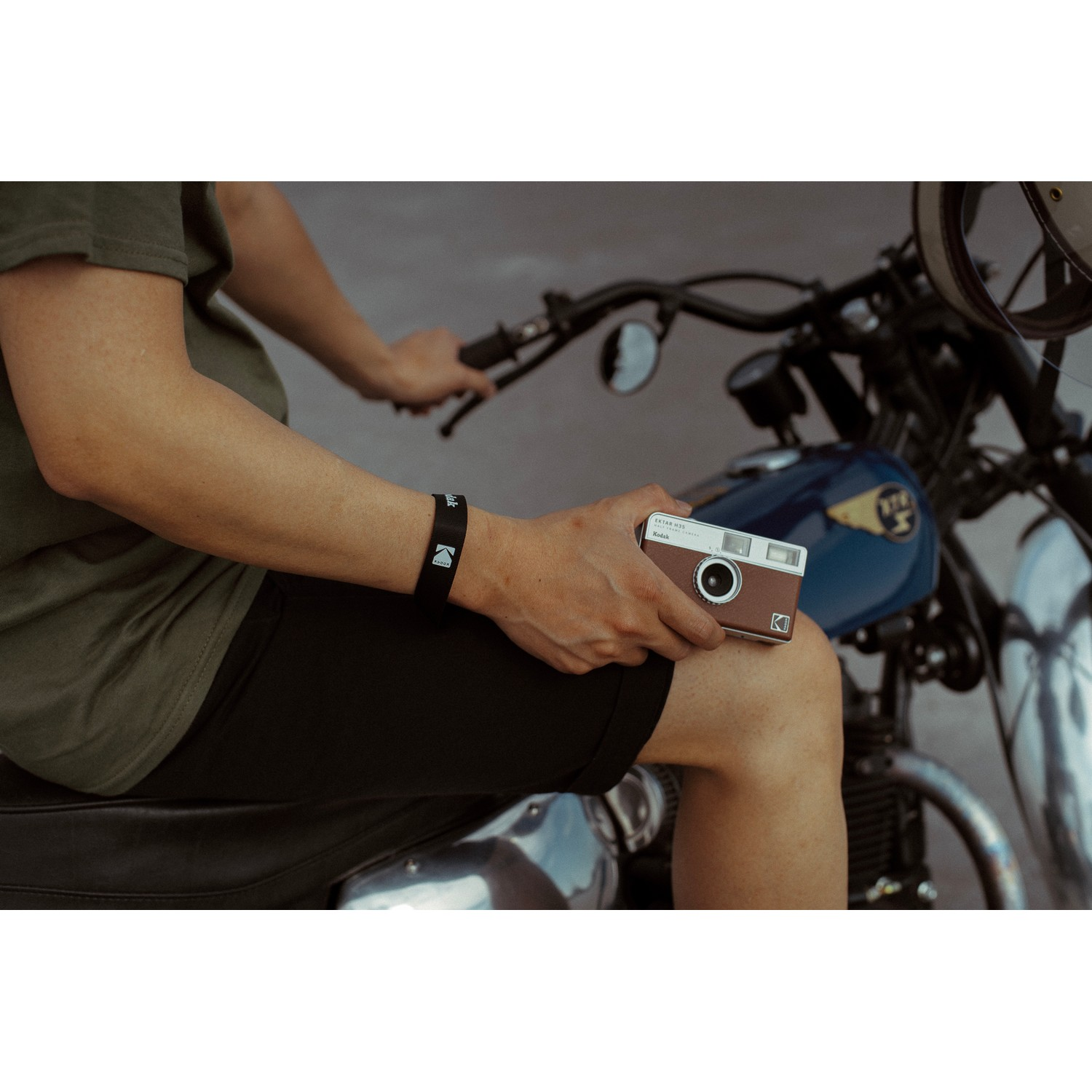 A person holding a brown camera. They're sitting on a motorcycle. A dark background.