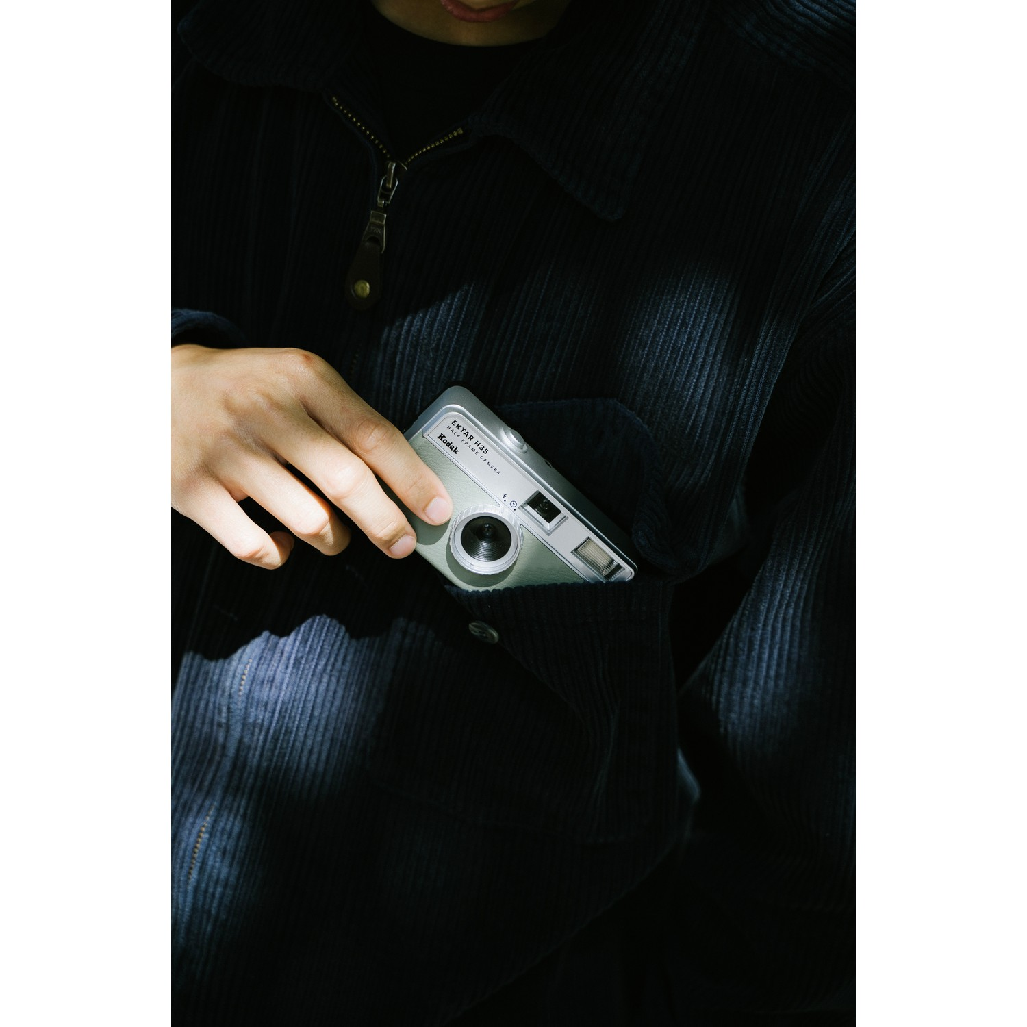 A person's hand holds a silver camera inside a pocket. The jacket is navy corduroy.