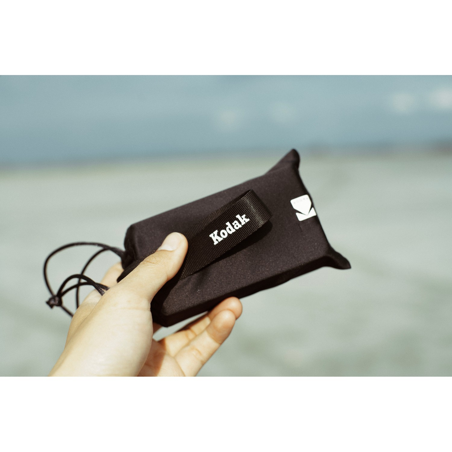 A hand holds a black pouch with the Kodak logo. The background is a blurry sky and water.