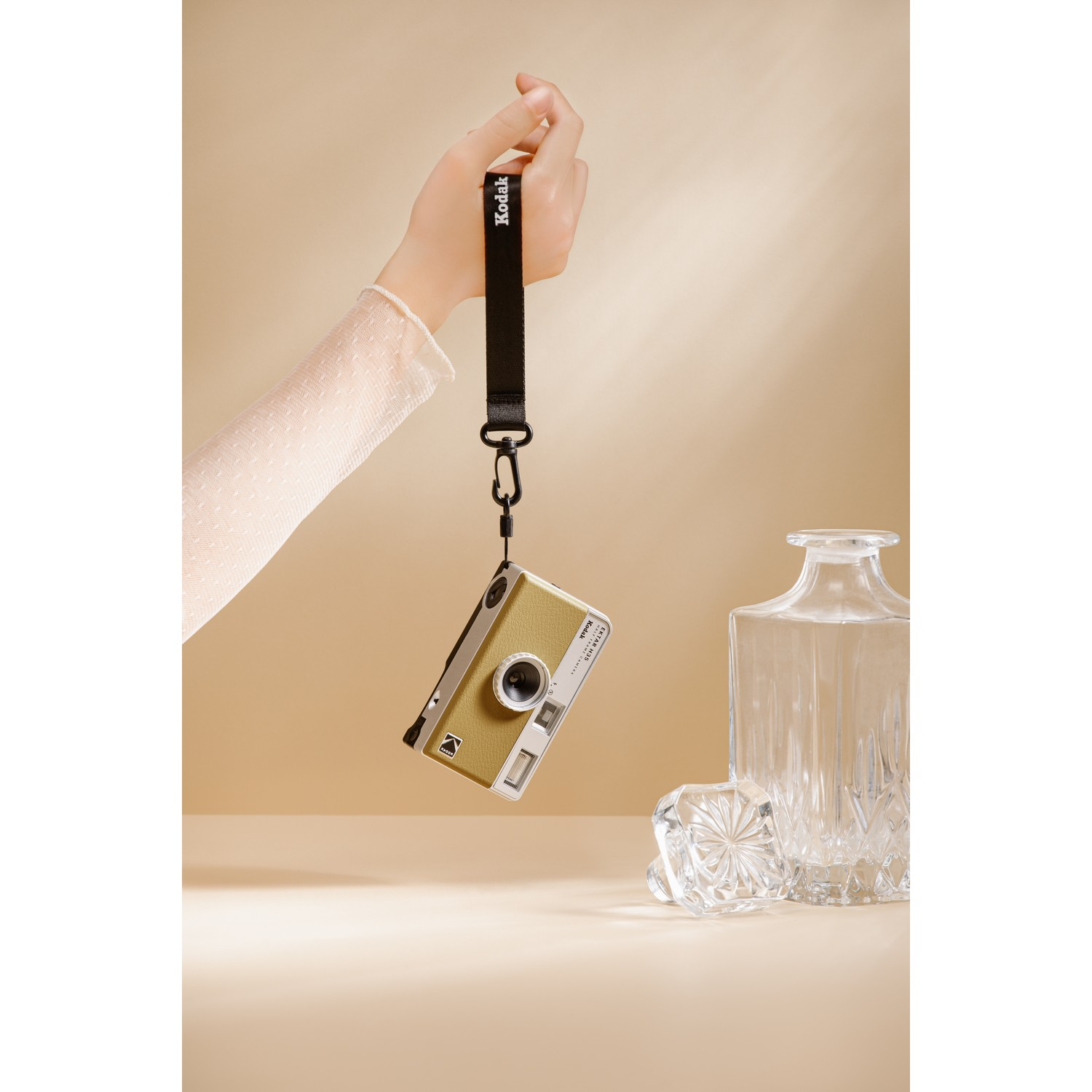 A hand holding a yellow and green Kodak camera hanging from a black strap near a glass vase.