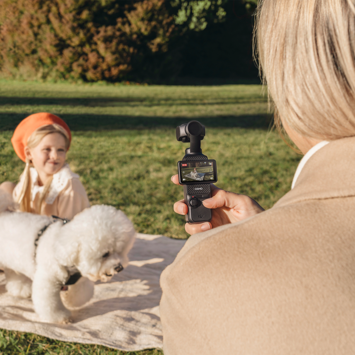 Una persona tiene in mano una piccola fotocamera, filmando un cane e una bambina sull'erba. È una giornata di sole, ambiente giardino.