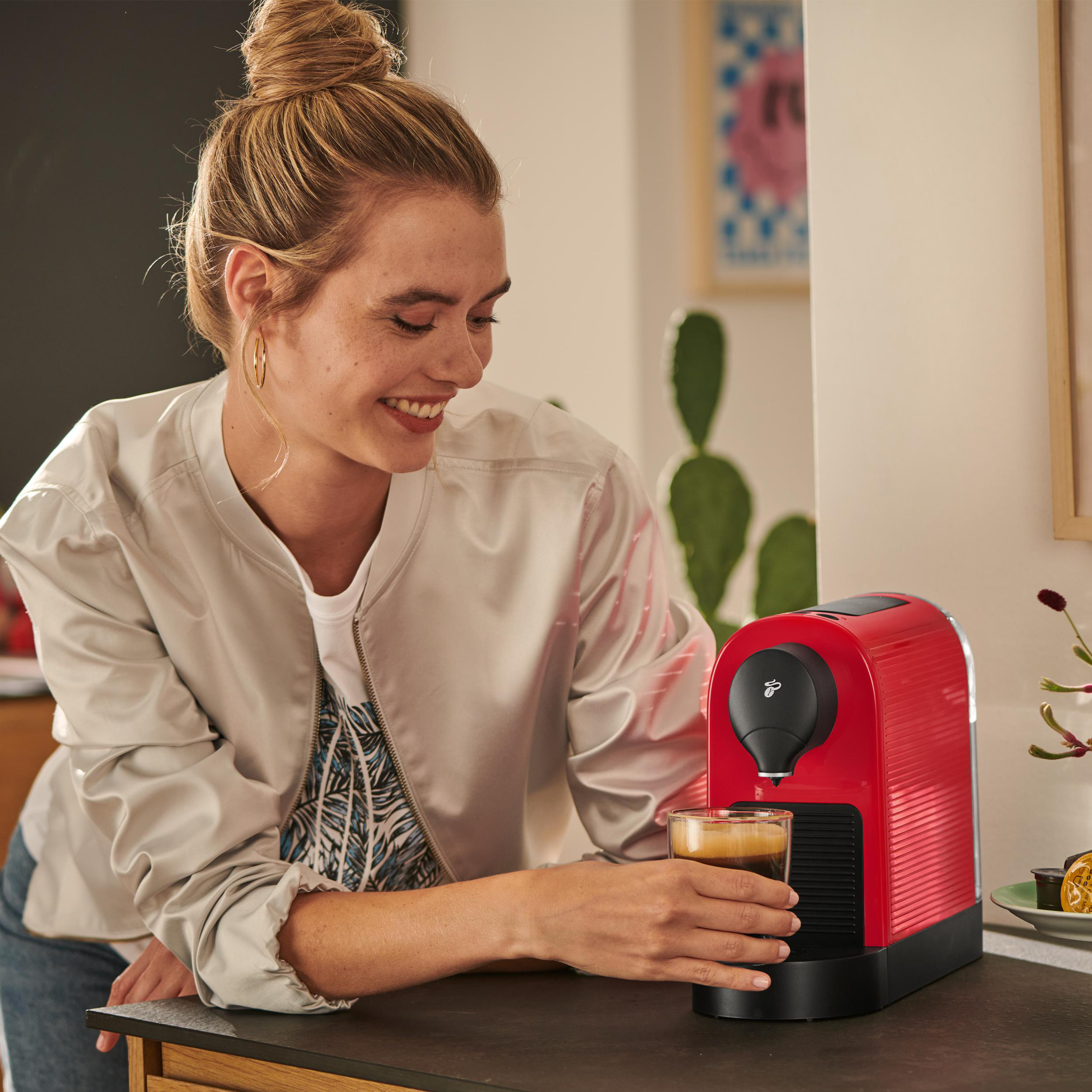 Eine Frau, die Kaffee mit einer roten Kaffeemaschine zubereitet, lächelt. Im Hintergrund ein Kaktus und Kunst.
