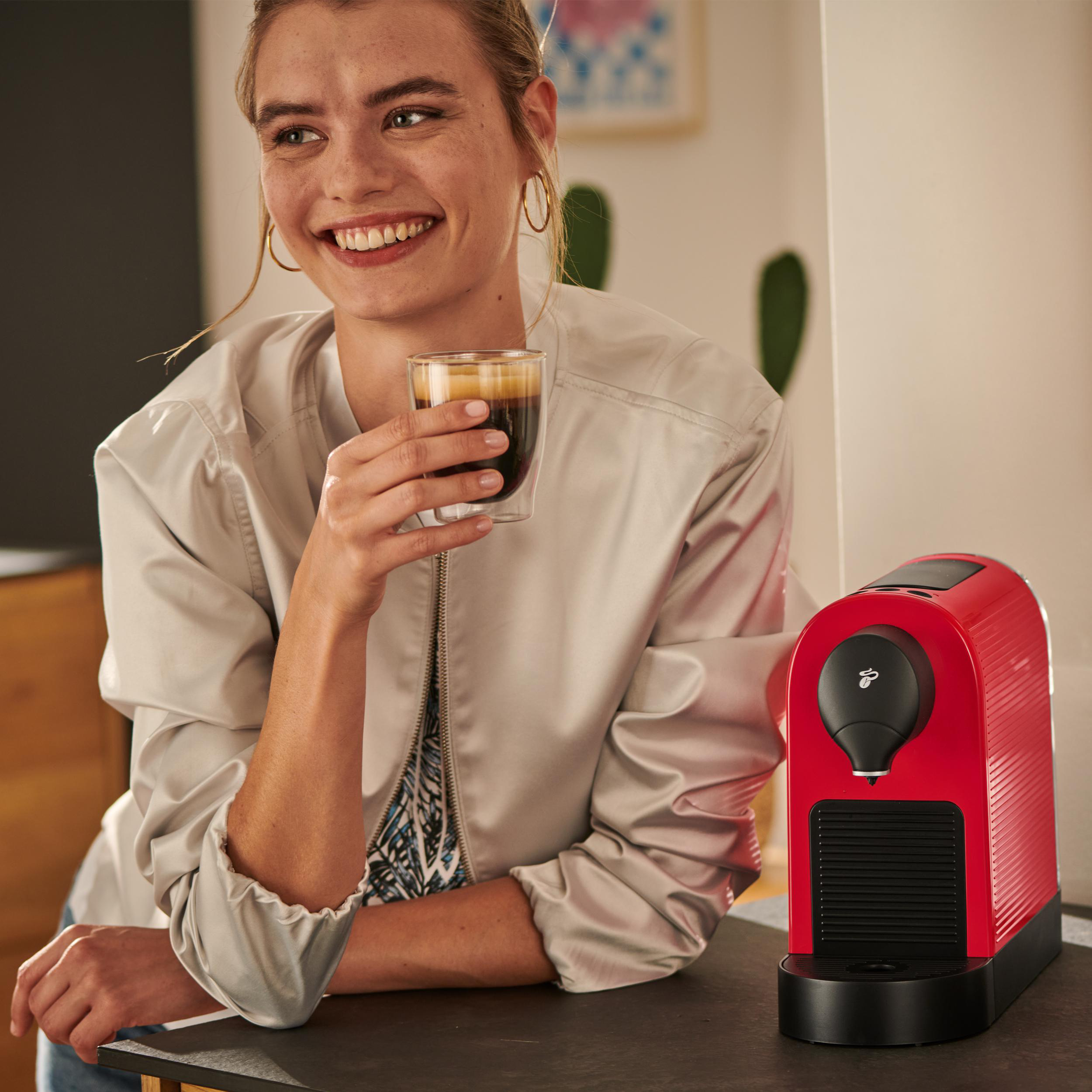 Lächelnde Frau, hält Kaffeeglas neben roter Kaffeemaschine auf Theke.