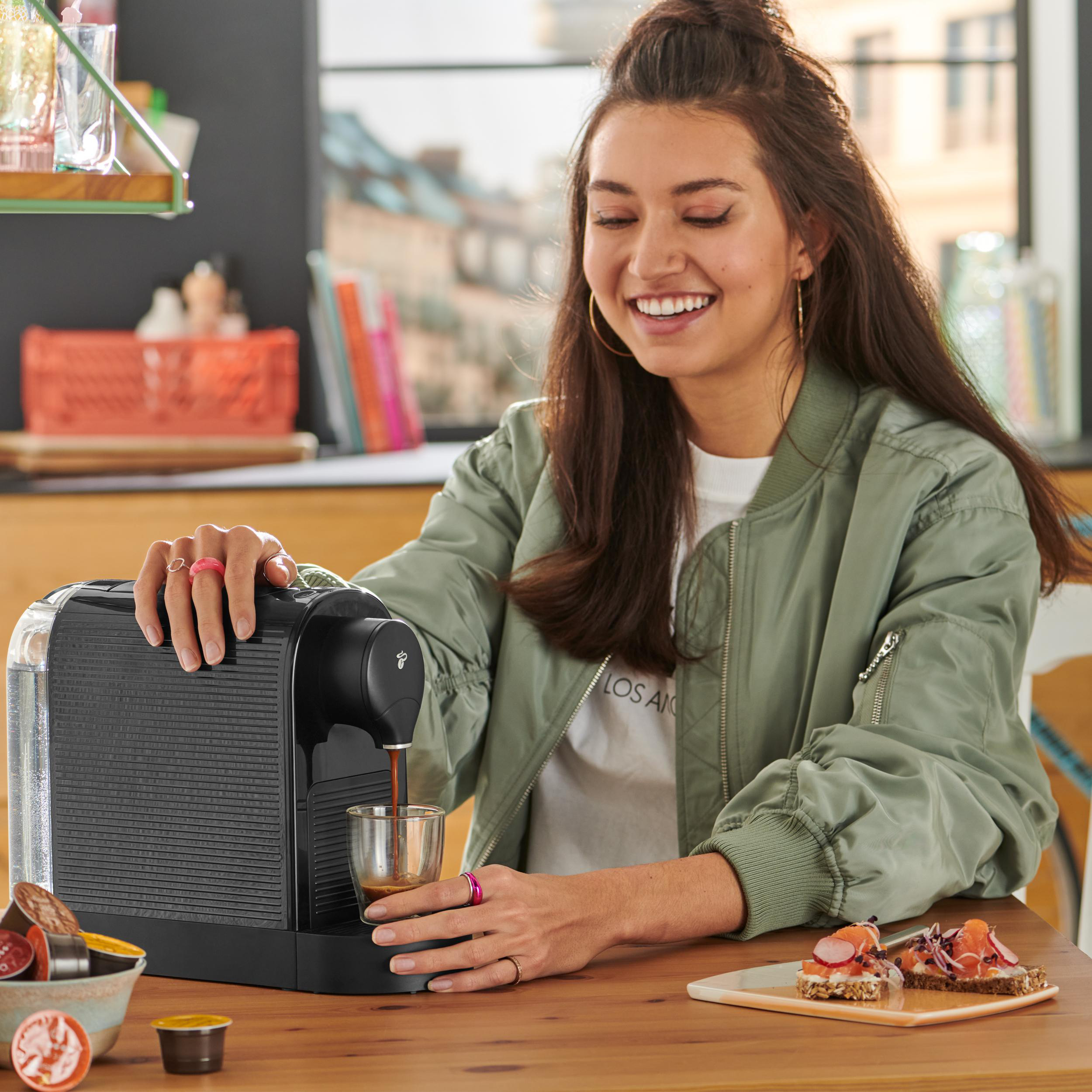 Frau bereitet Kaffee mit einer schwarzen Espressomaschine zu. Sie lächelt und hält eine Tasse. Essen auf dem Tisch.