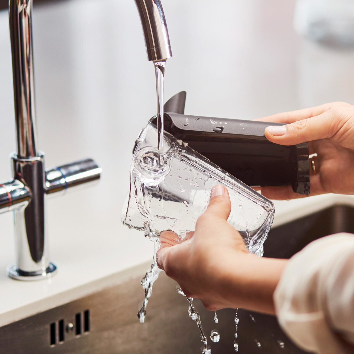 Ręce osoby napełniają przezroczysty szklany pojemnik wodą z srebrnej baterii w zlewie.