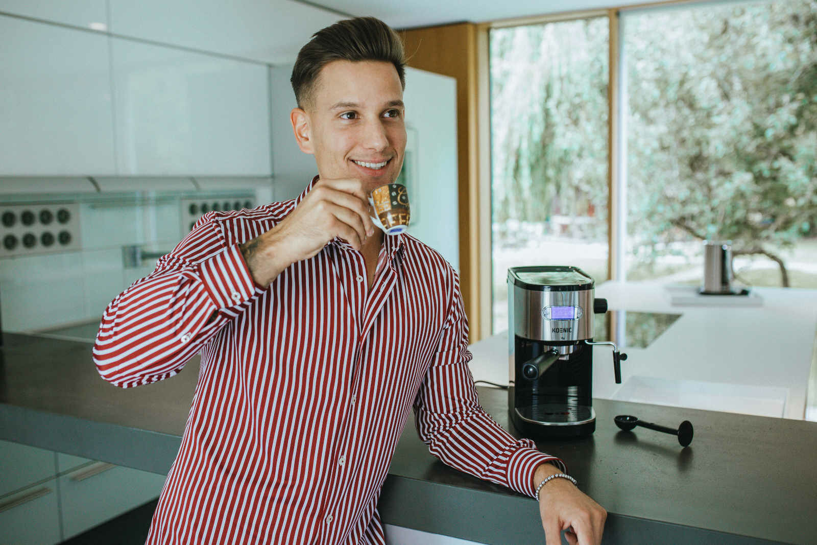 Mężczyzna w czerwono-białą koszulę w paski uśmiecha się, trzymając filiżankę kawy w pobliżu ekspresu do kawy.