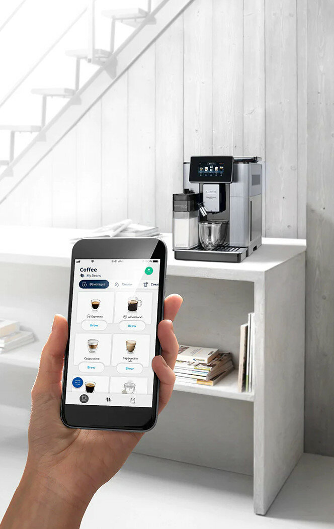 Ręka trzyma telefon, ekspres do kawy na półce. Widoczny ekran aplikacji, nowoczesna kuchnia.