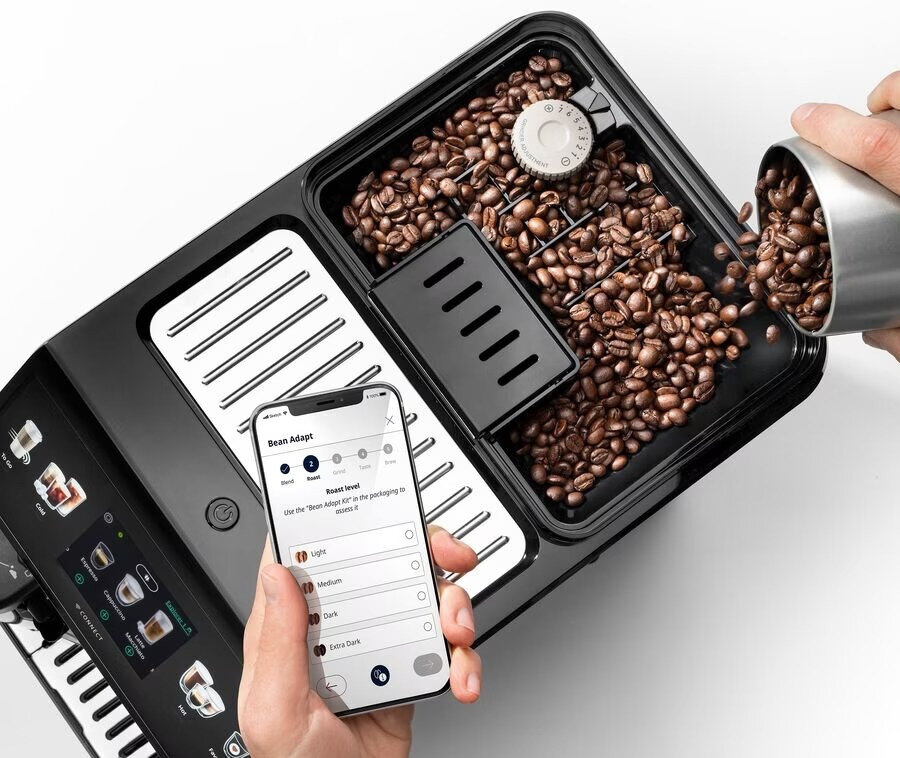 Ekspres do kawy z ziarnami, ręka wsypuje ziarna, telefon pokazuje ustawienia.