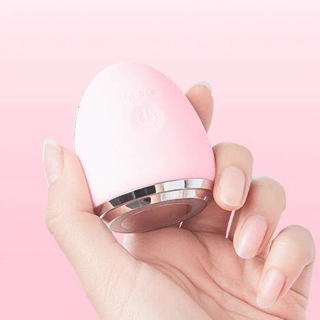 Ręka trzymająca różowe urządzenie do oczyszczania twarzy na różowym tle. Urządzenie ma srebrną obwódkę.