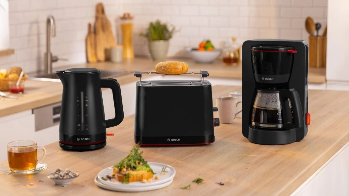 Blat kuchenny z czarnym czajnikiem, tosterem z bajglem i ekspresem do kawy. Jest talerz z jedzeniem.