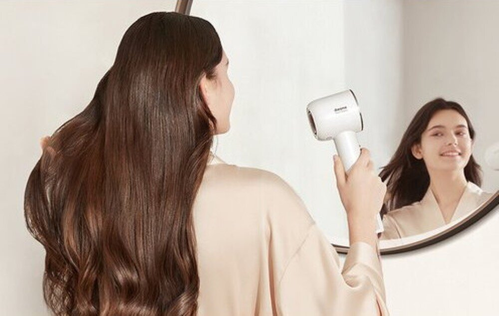 Kobieta z długimi włosami suszy włosy białą suszarką, patrząc w lustro.