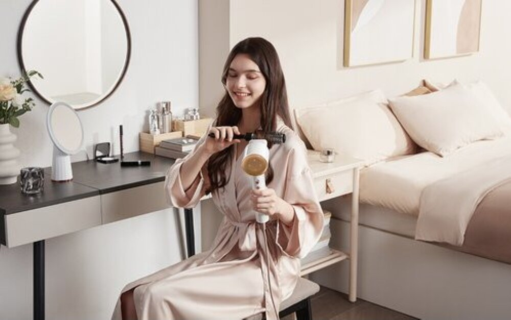 Kobieta używa suszarki do włosów i grzebienia. Uśmiecha się. Lustro, toaletka i łóżko w tle.